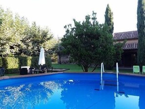 Une piscine avec un escalier, entourée d’arbres et dotée d’un espace patio avec des chaises et un parasol.