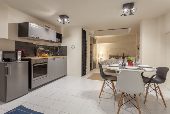 A modern kitchen with stainless steel appliances, a dining area with a round table and chairs, and a visible bed in the adjacent room.