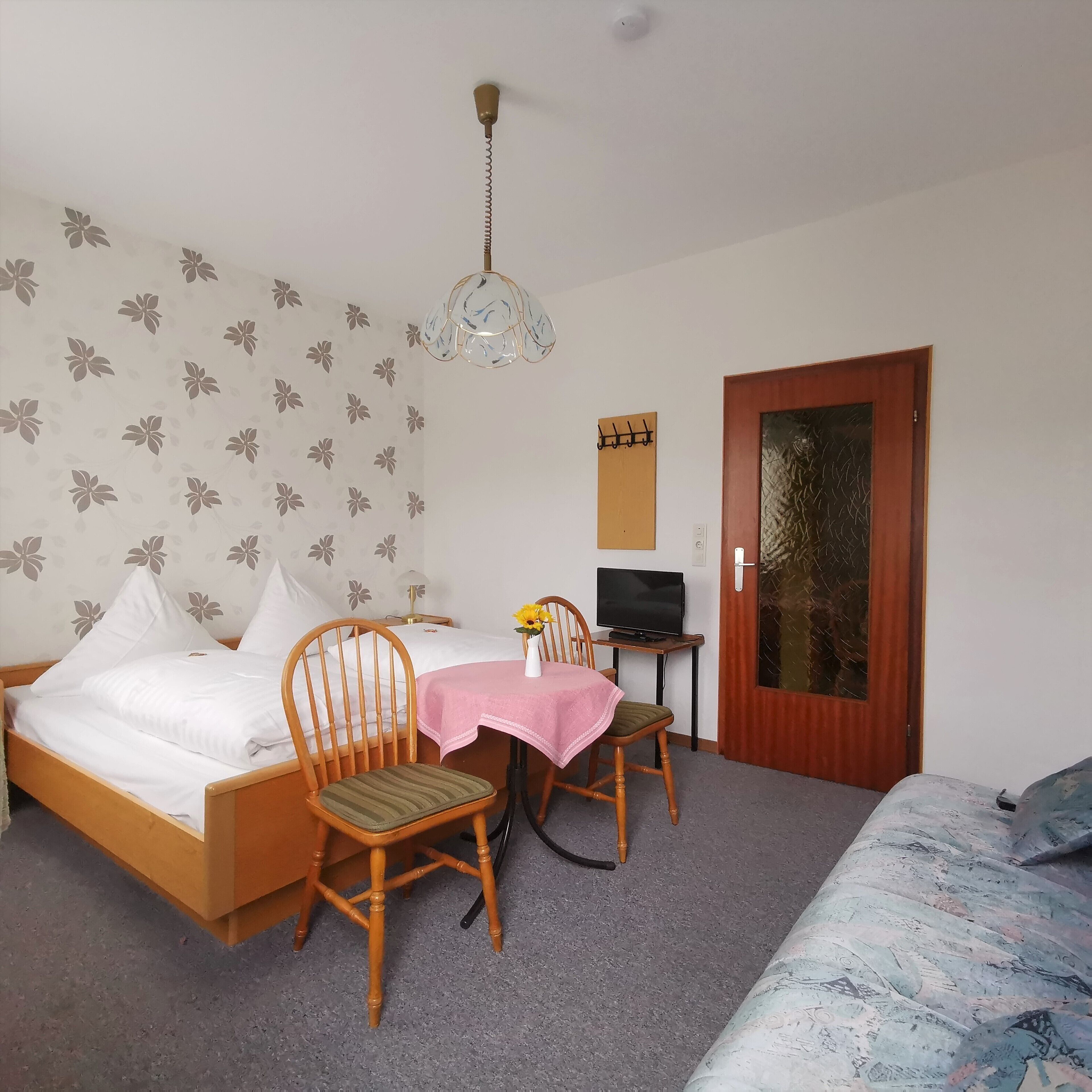 Ein Schlafzimmer mit einem Bett, zwei Holzstühlen, einem kleinen Tisch mit rosa Tischdecke, einem Fernseher auf einem Schrank und einer Tür mit Fensterteil.