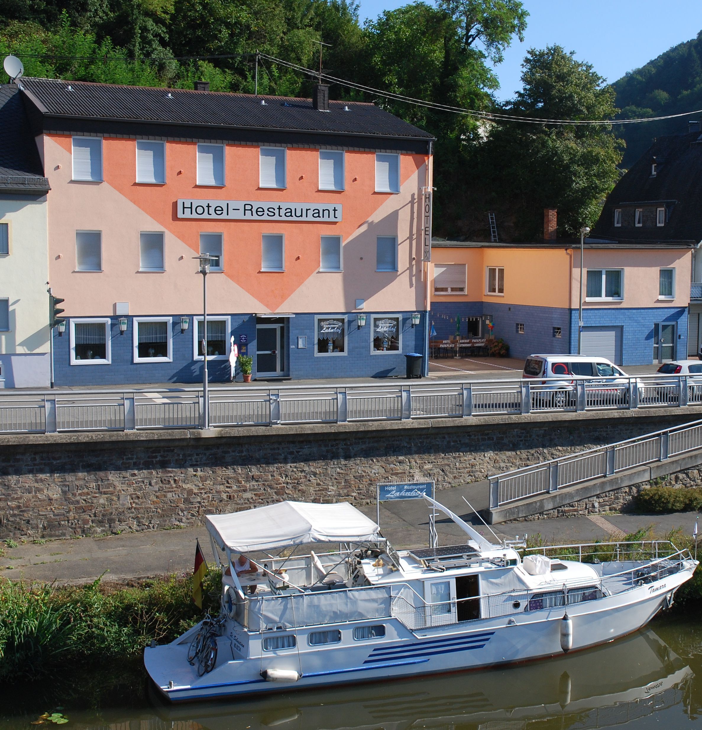 Ein Hotel mit der Aufschrift 'Hotel-Restaurant' an der Fassade, einem vor der Tür liegenden Boot und einer Fußgängerbrücke.