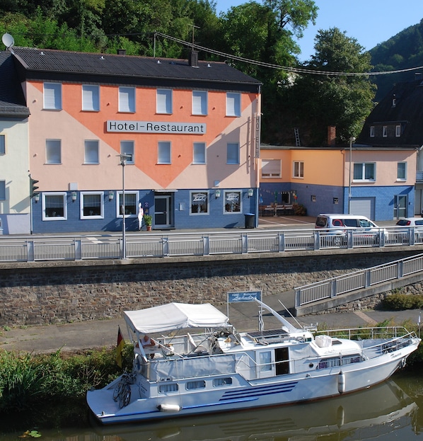 A hotel with a sign reading "Hotel-Restaurant" on the facade, a boat docked in front, and a pedestrian bridge.