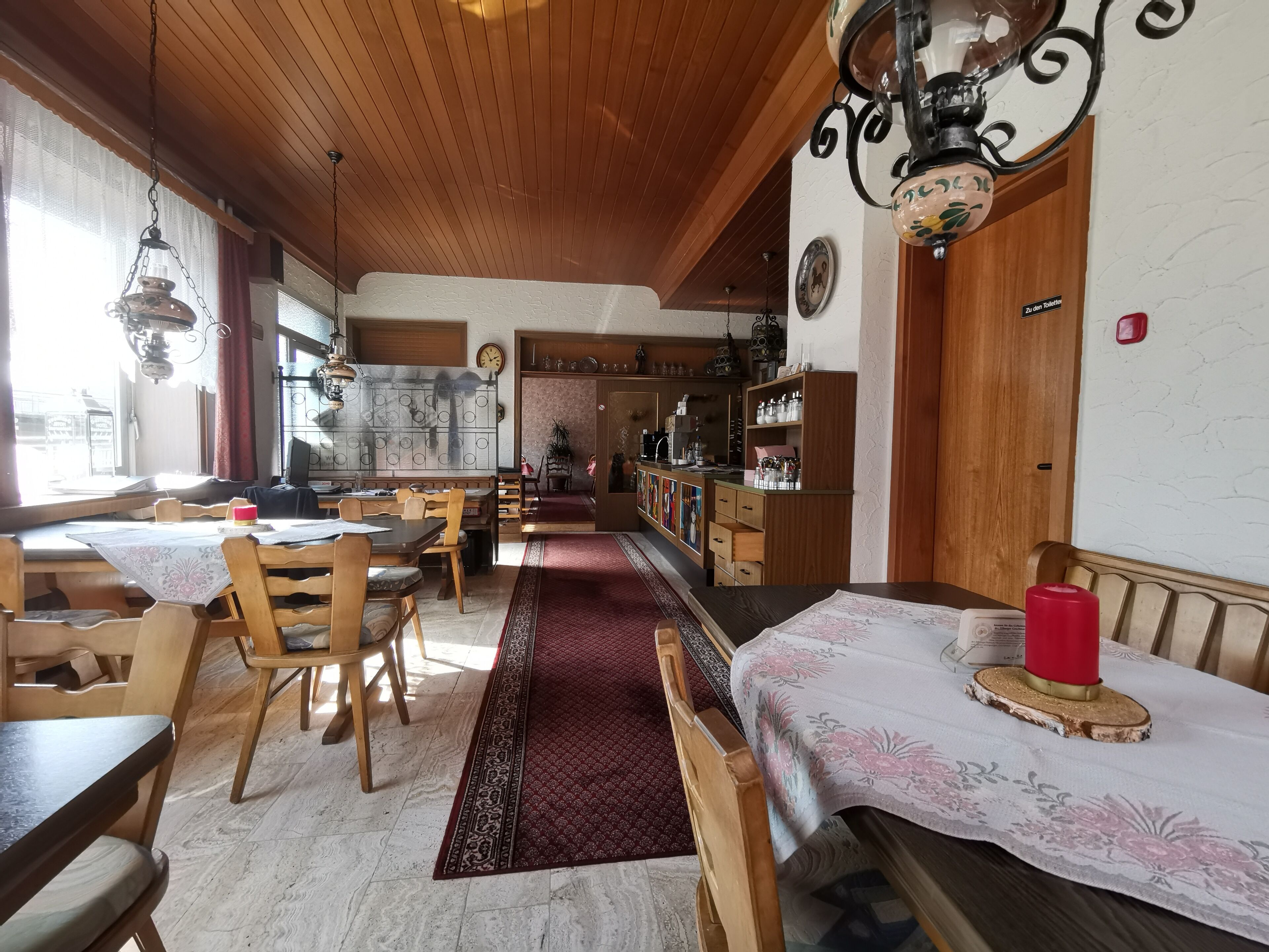 Un restaurant chaleureux avec des tables et des chaises en bois, une moquette rouge et une cuisine visible au fond de la salle.