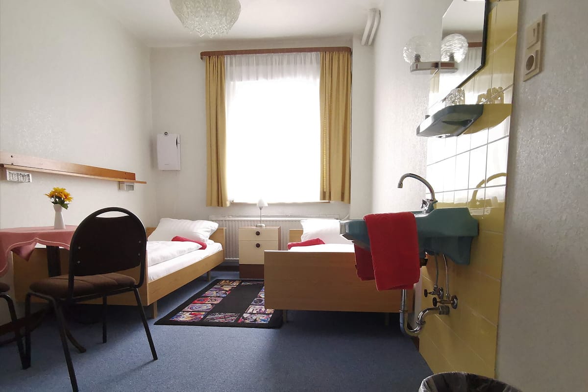 Ein kompaktes Hotelzimmer mit einem Einzelbett, einem kleinen Schreibtisch, einem Stuhl, einem Fenster mit Vorhängen, einem Waschbecken und einem Mülleimer.