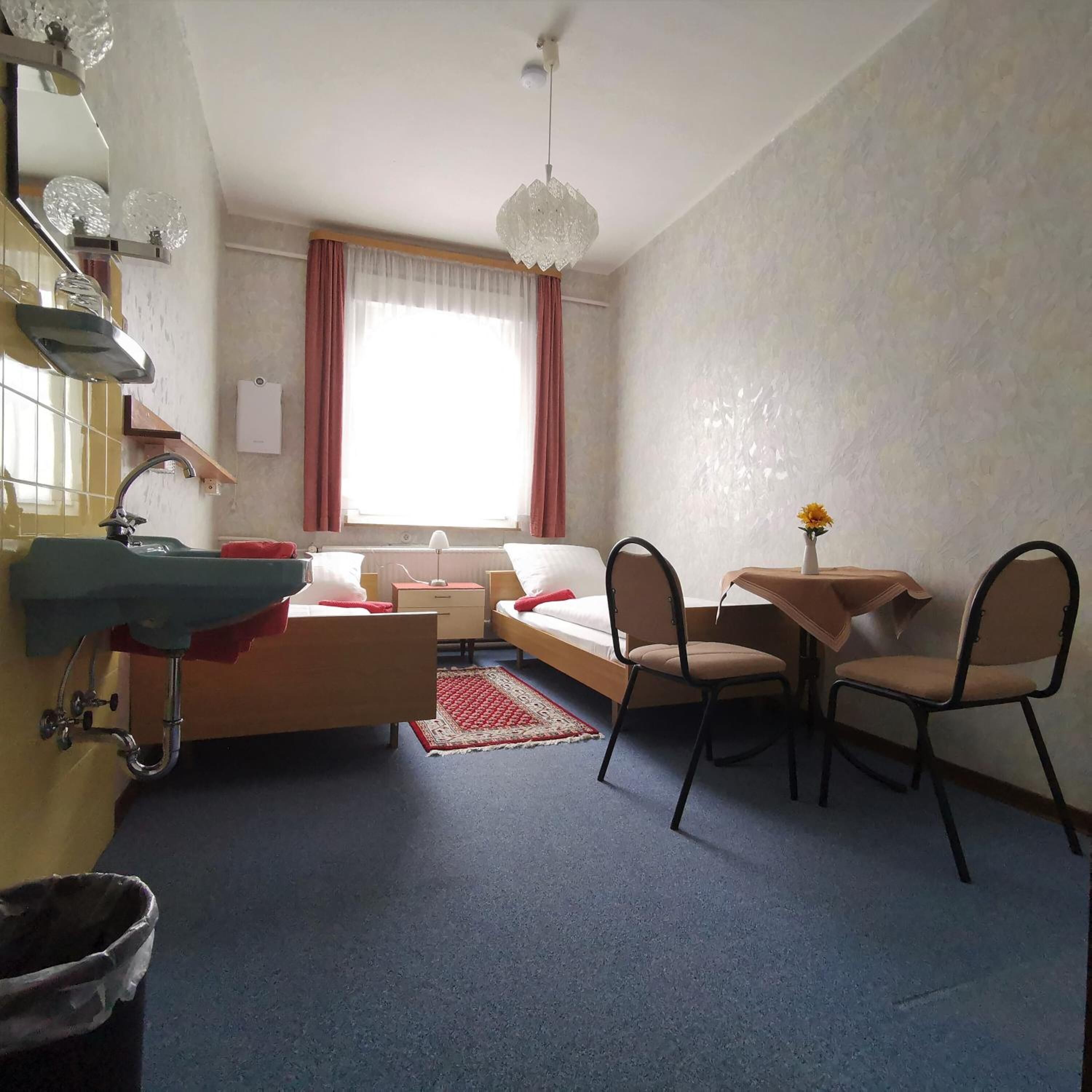 Une chambre d’hôtel de taille réduite, comprenant un lit, une petite table avec des chaises, un lavabo et une fenêtre avec des rideaux.