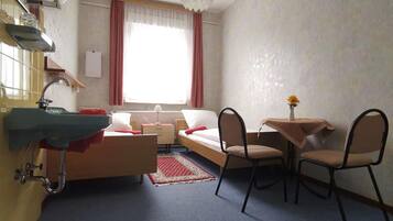 Une chambre d’hôtel de taille réduite, comprenant un lit, une petite table avec des chaises, un lavabo et une fenêtre avec des rideaux.