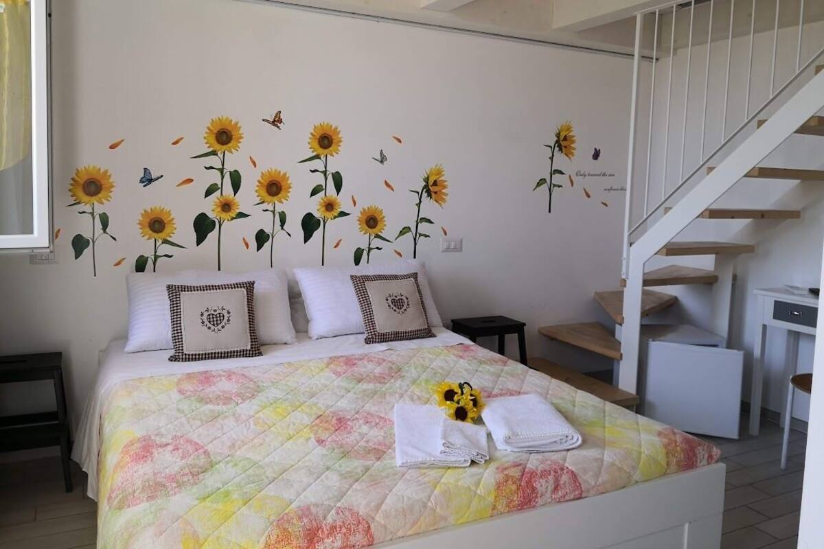 A bedroom with a bed, a wall mural of sunflowers, a staircase, and a small desk.