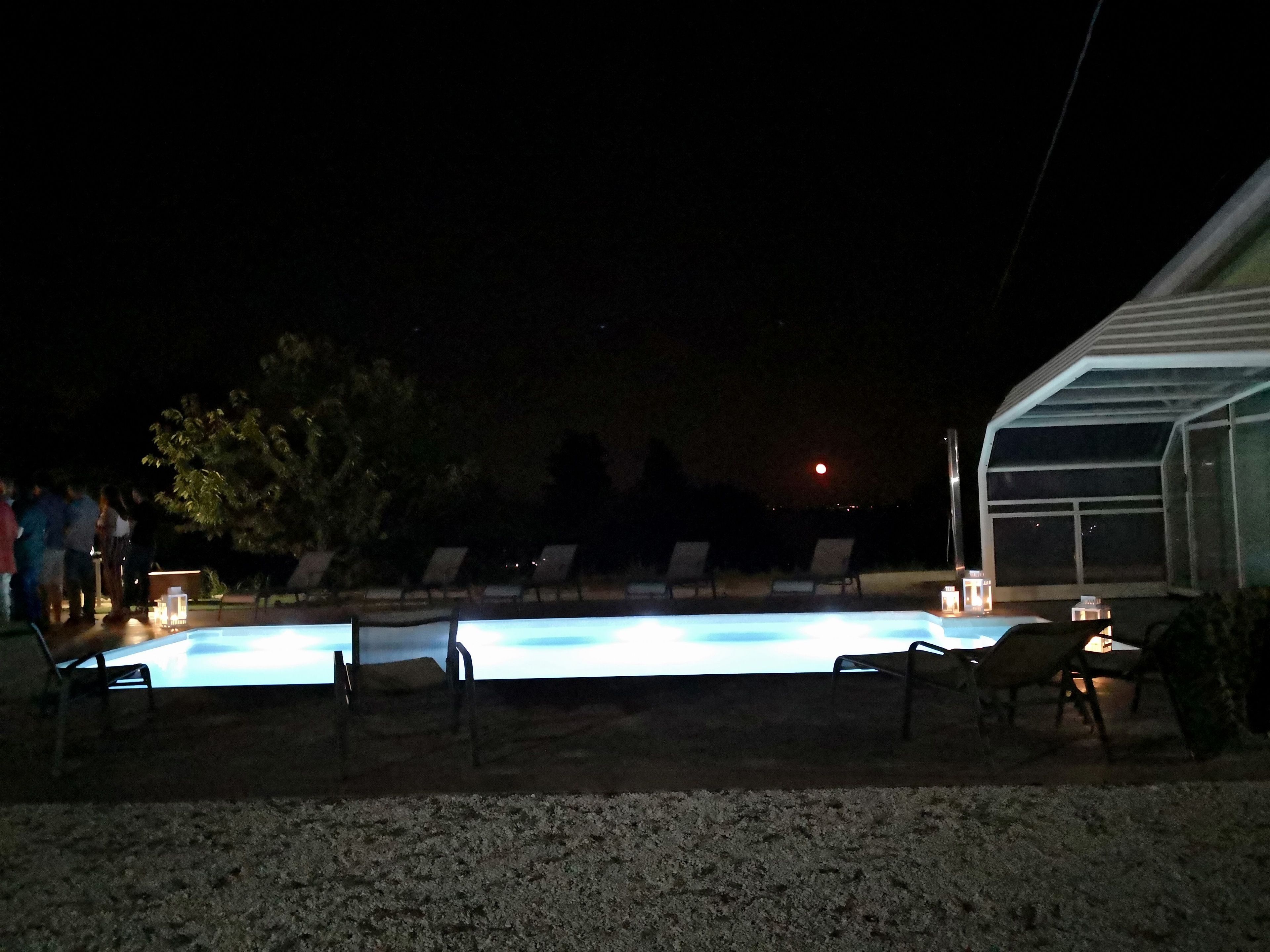 Una piscina con sillones de descanso y una zona cubierta, rodeada de árboles y un edificio.