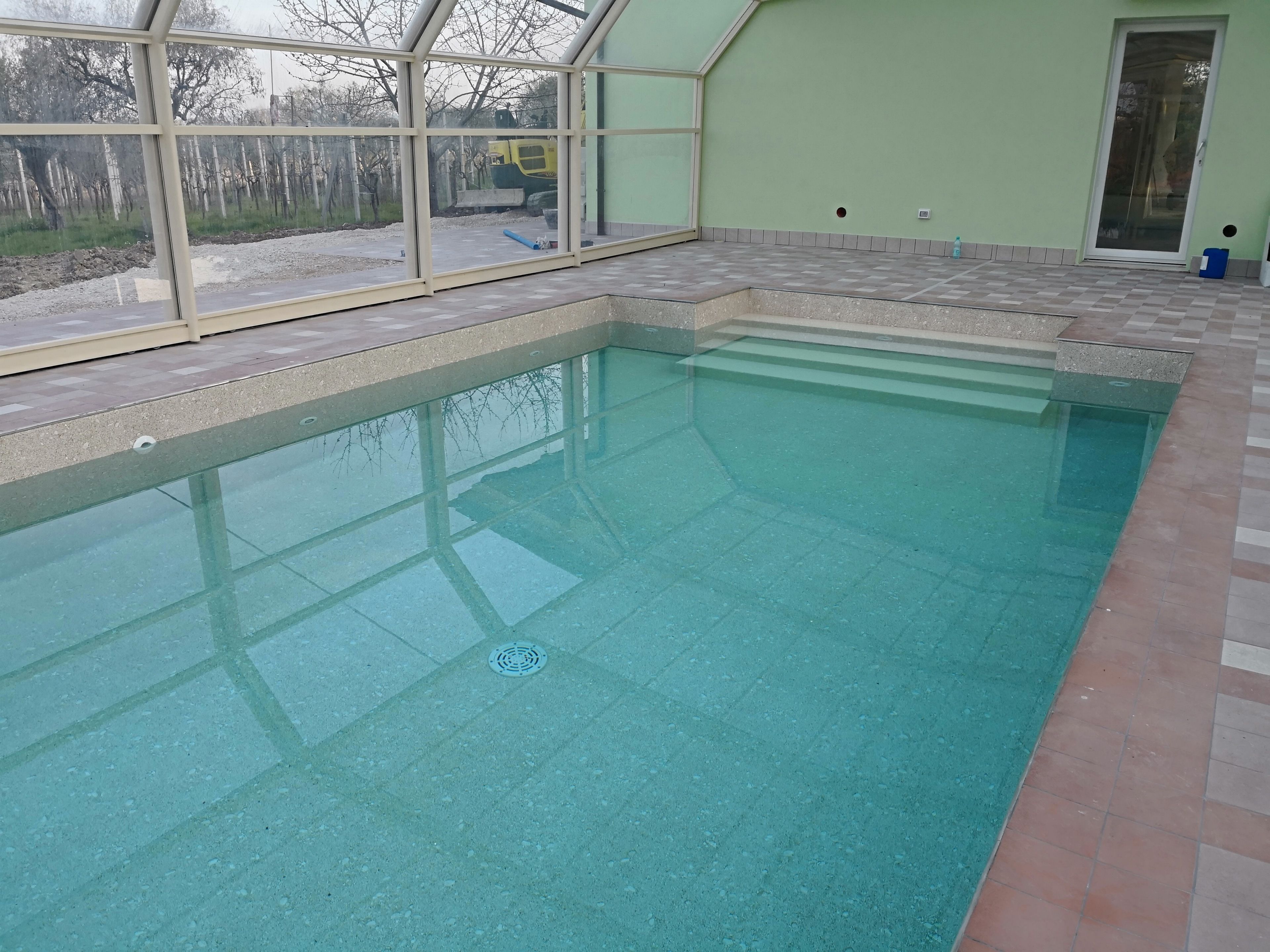 An indoor swimming pool with clear water, surrounded by a tiled deck and a glass enclosure.