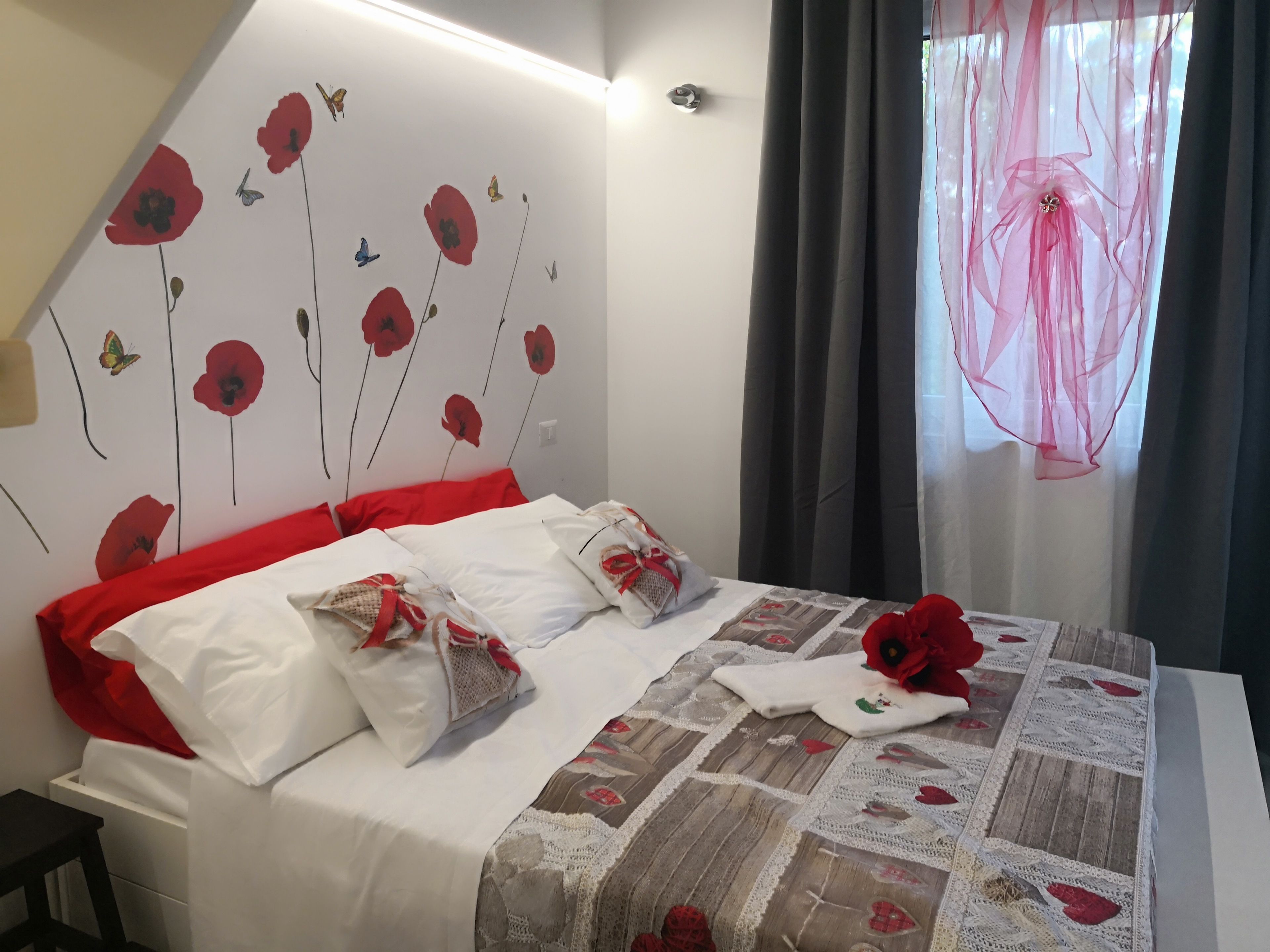Una cama bien hecha con ropa de cama blanca y roja, un colcha con motivos florales y una pared decorada con flores y mariposas rojas.