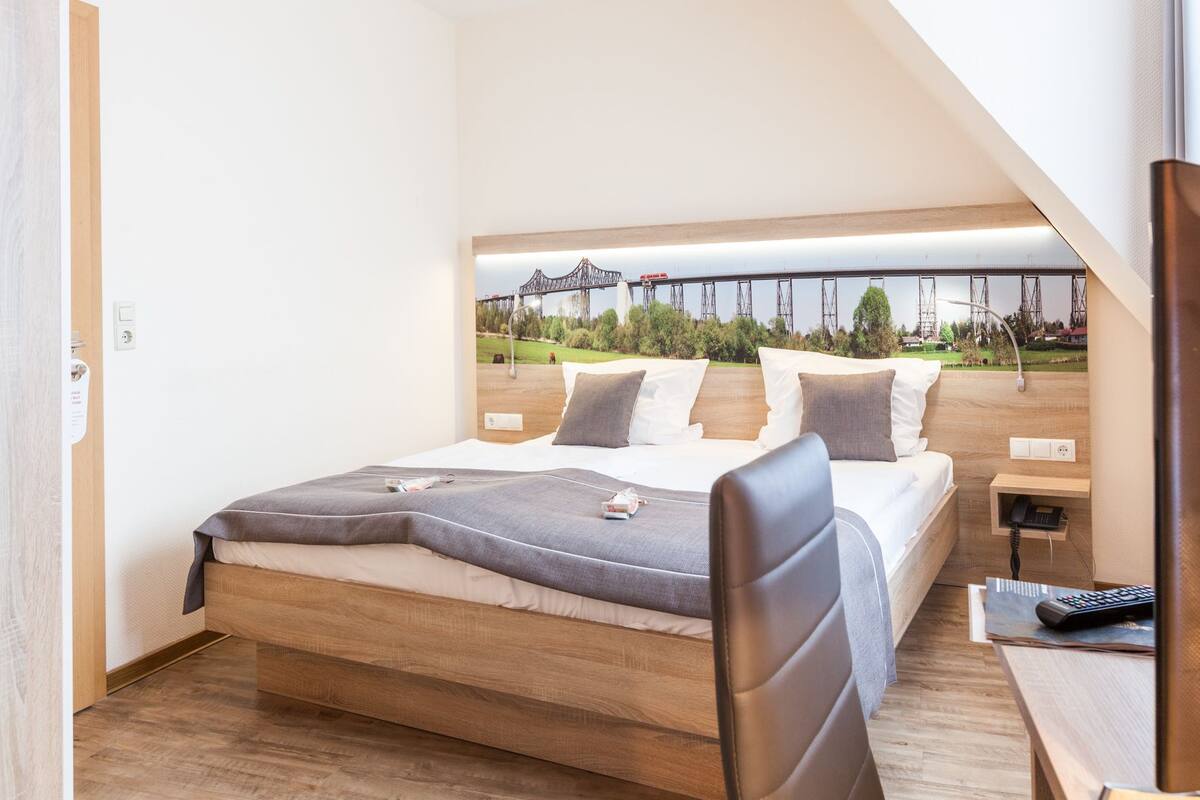 Ein Schlafzimmer mit einem Bett, einem Schreibtisch und einem Stuhl. Ein Fenster bietet Blick auf eine Brücke und Bäume.