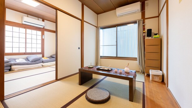 Une pièce de style japonais traditionnel, avec un sol recouvert de tatamis, une basse table en bois ornée d’objets utilisés lors des cérémonies du thé, une porte coulissante et une fenêtre avec des stores.