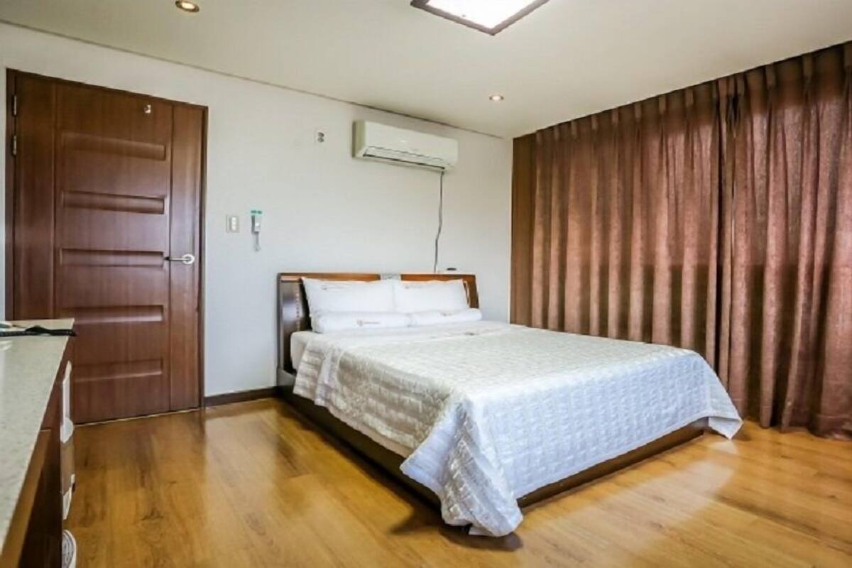 Habitación de hotel con piso de madera, cama con ropa blanca, puerta de madera y cortina marrón.