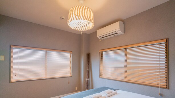 A bedroom with two large windows with wooden blinds, a bed with a blue blanket, and an air conditioning unit on the wall. - YUYU House - Kohama Mei (Osaka)