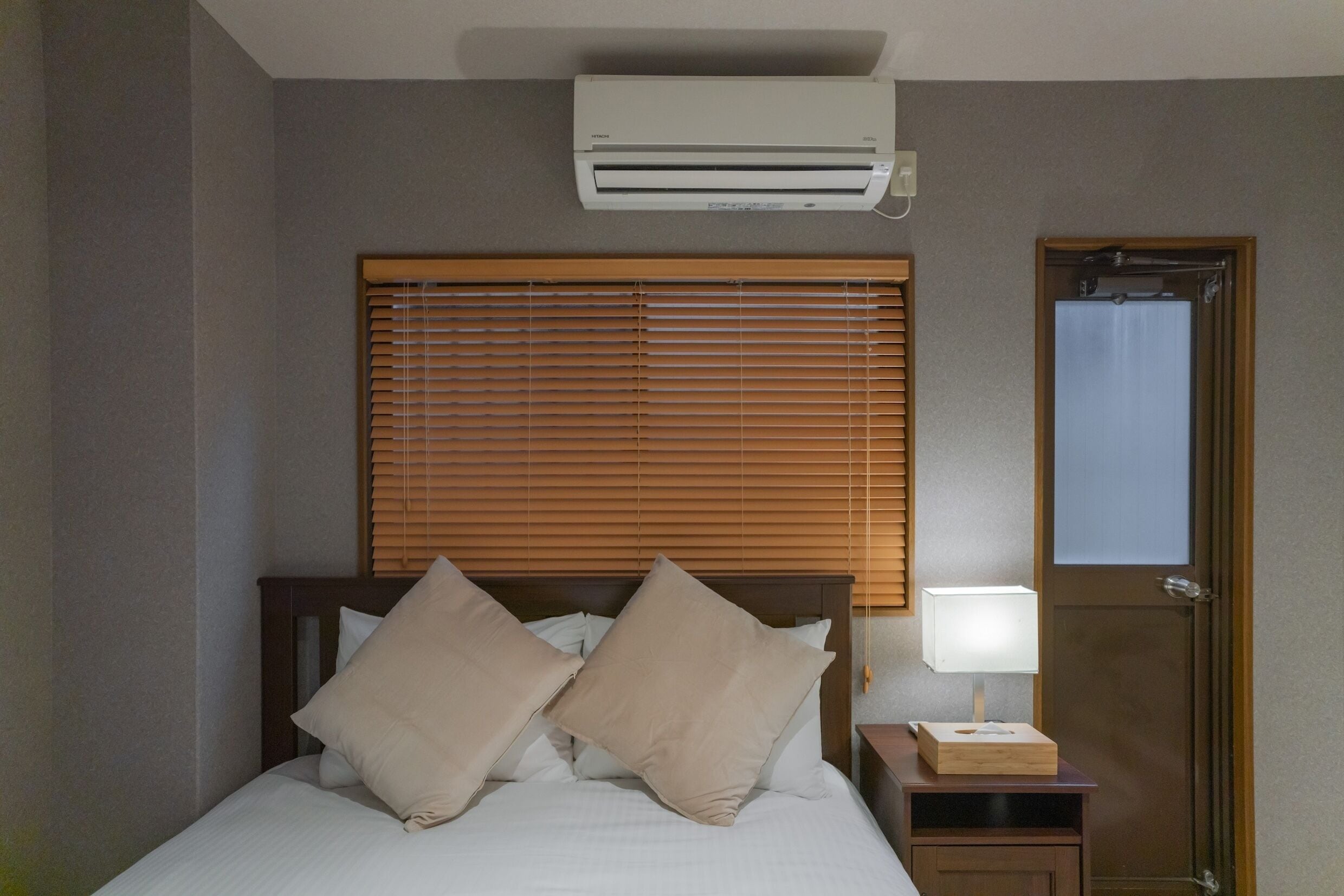 Quarto de hotel com cama, mesa de cabeceira, abajur, janela com persianas e ar condicionado.