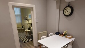A dining area with a white table and chairs, a clock on the wall, and a view into another room with a kitchen and a striped rug. - First - Slateford House Apartment (Edinburgh)
