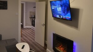 A living room with a flat-screen TV mounted on the wall, a fireplace with a visible fire, a striped rug, and a modern pendant light. - First - Slateford House Apartment (Edinburgh)
