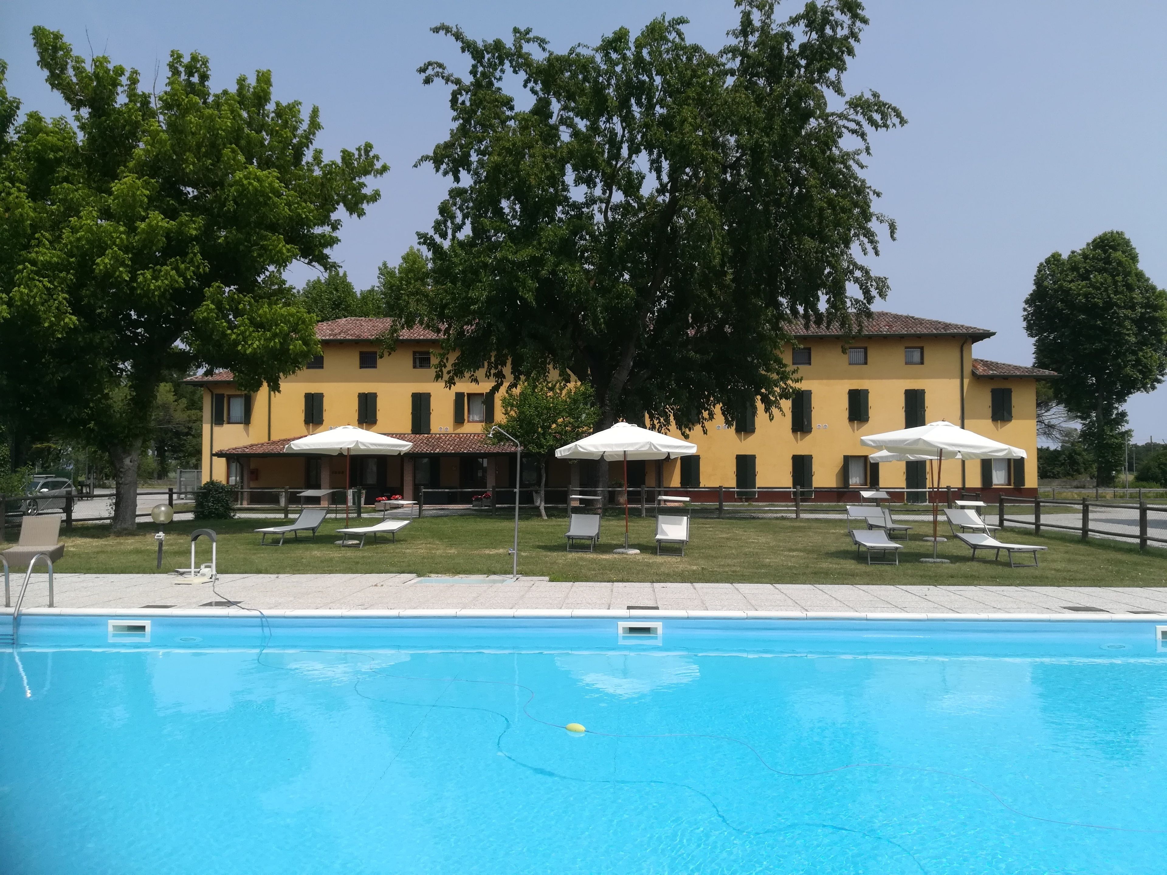 A swimming pool with lounge chairs and umbrellas, a yellow building with multiple windows, and trees in the background.