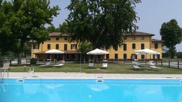 A swimming pool with lounge chairs and umbrellas, a yellow building with multiple windows, and trees in the background.