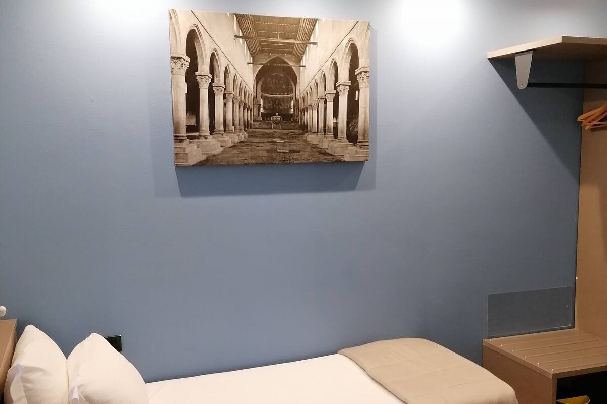 Een kleine kamer met een enkel bed, een houten vloer, beddengoed in het wit met twee kussens, een klein nachtkastje van hout en een foto aan de muur van een architectonisch gangpad.