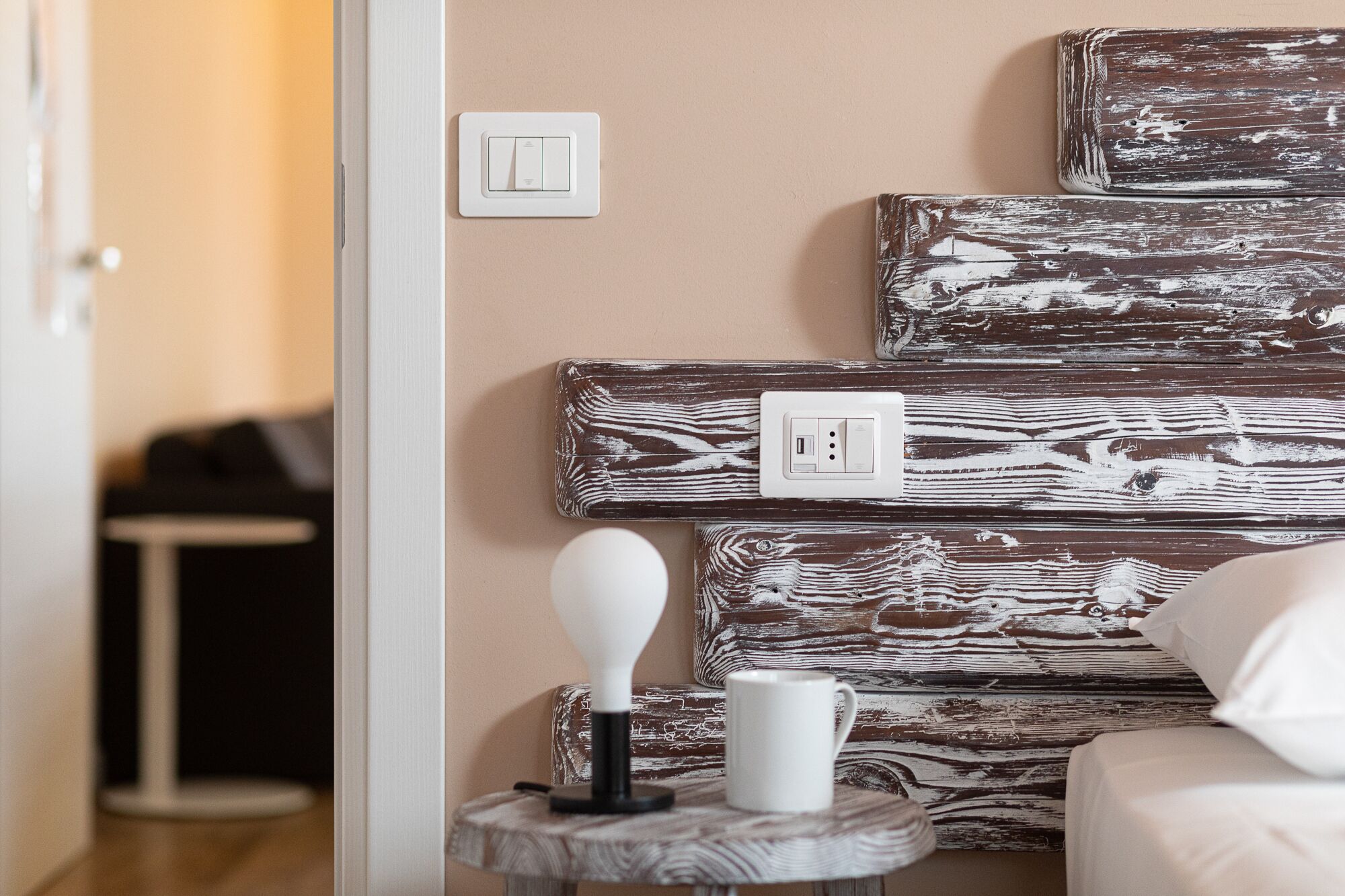A room with a bed, a bedside table with a lamp and a mug, a wall-mounted power outlet, and a door leading to another room.