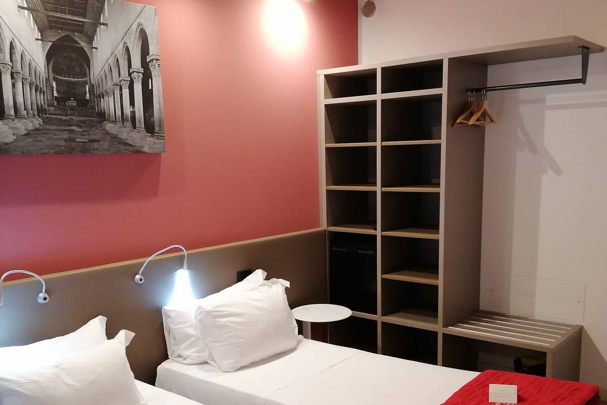 Hotelkamer met twee bedden, elk met een rode deken en een wit kussen, een klein rond tafeltje, en een wandplank met een kapstok.