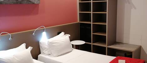 A hotel room with two beds, a red blanket on each, a white pillow on each bed, a small round table, and a wall-mounted shelf with a hanger.