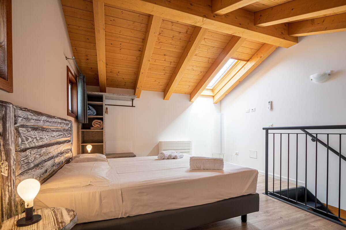 Een slaapkamer met een houten plafond, een bed met witte lakens, een bedlampje en uitzicht op een trap.