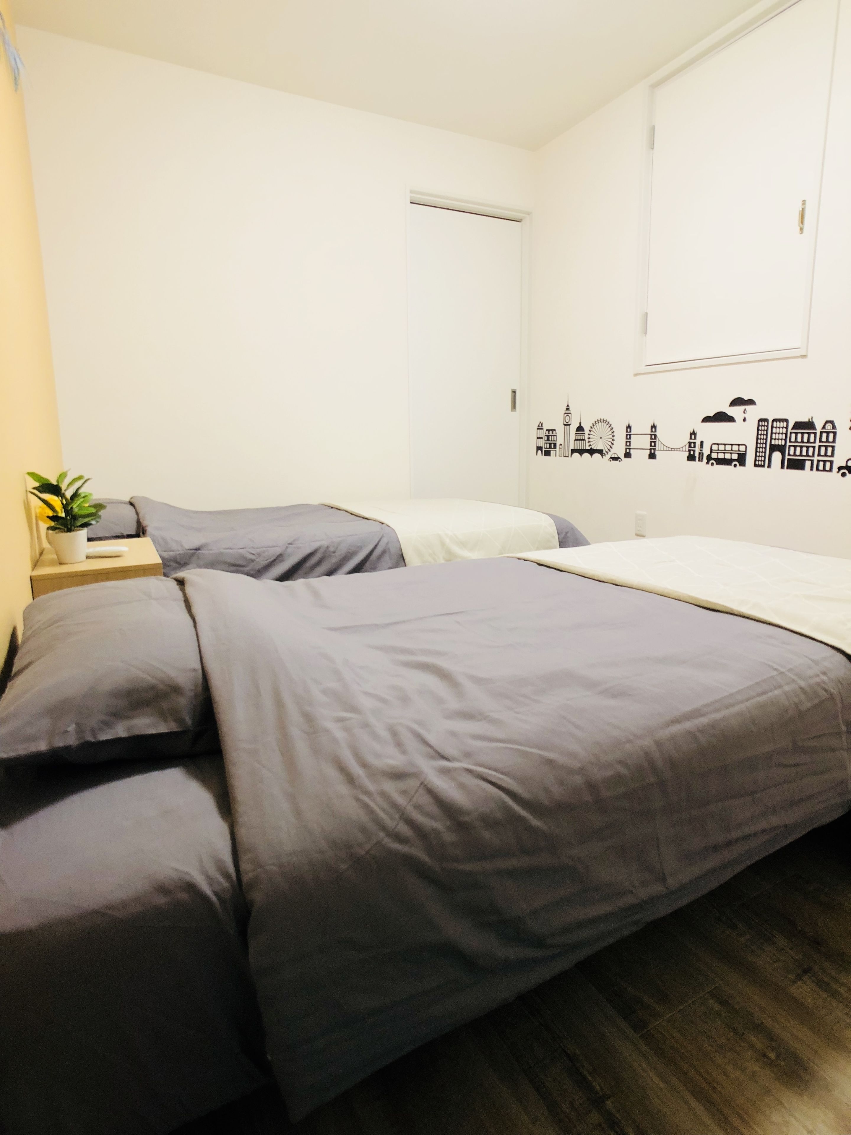 Une chambre avec deux lits, une petite table sur laquelle est posée une plante, et un mur décoré d’autocollants représentant des paysages urbains.