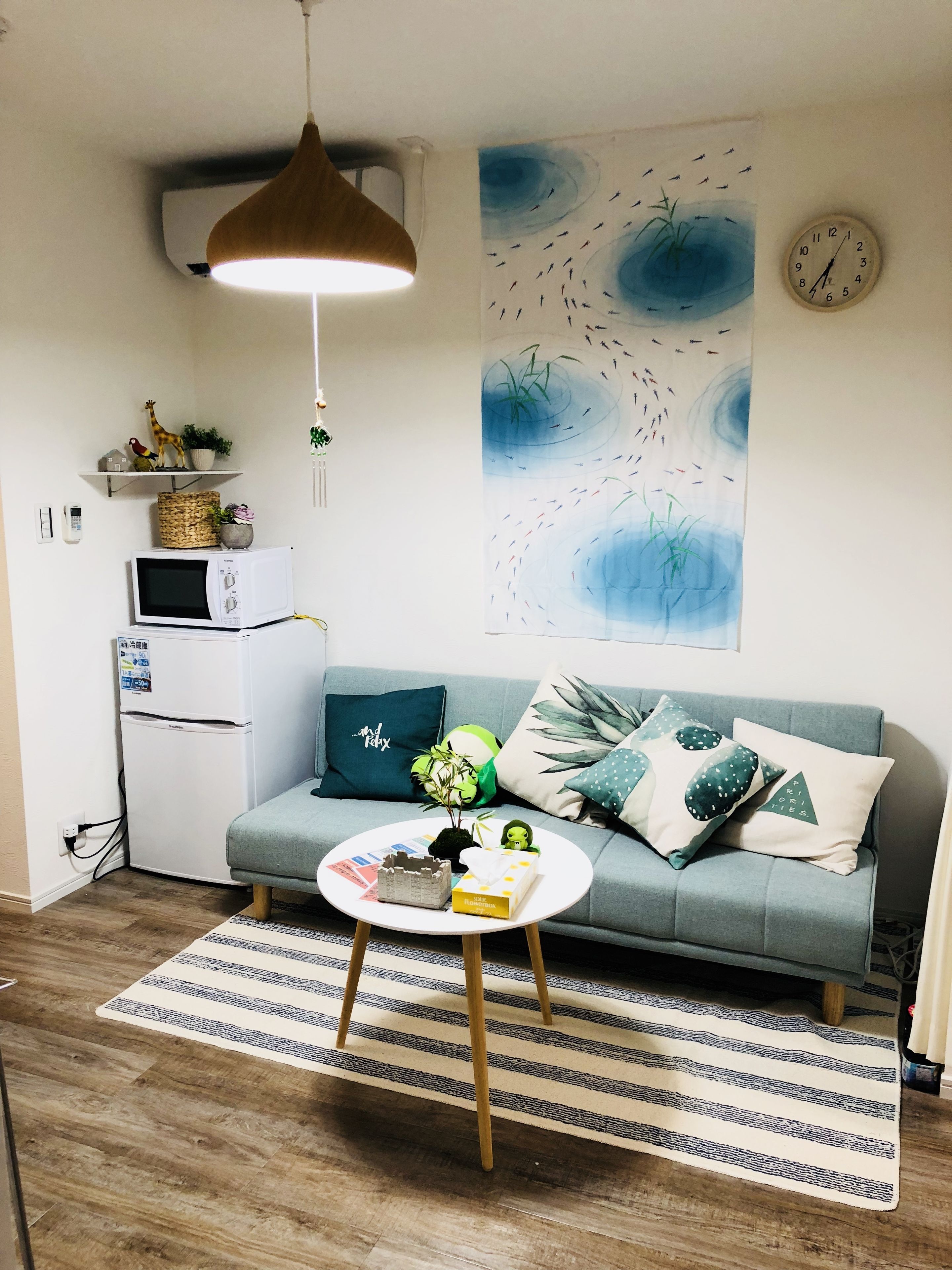 A cozy living room with a grey sofa, a round coffee table, a hanging pendant light, a wall clock, and a large abstract painting.