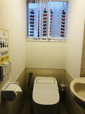 Une petite salle de bain avec des toilettes, un lavabo et une fenêtre agrémentée de rideaux colorés. Un panneau avec des instructions est affiché sur le mur.