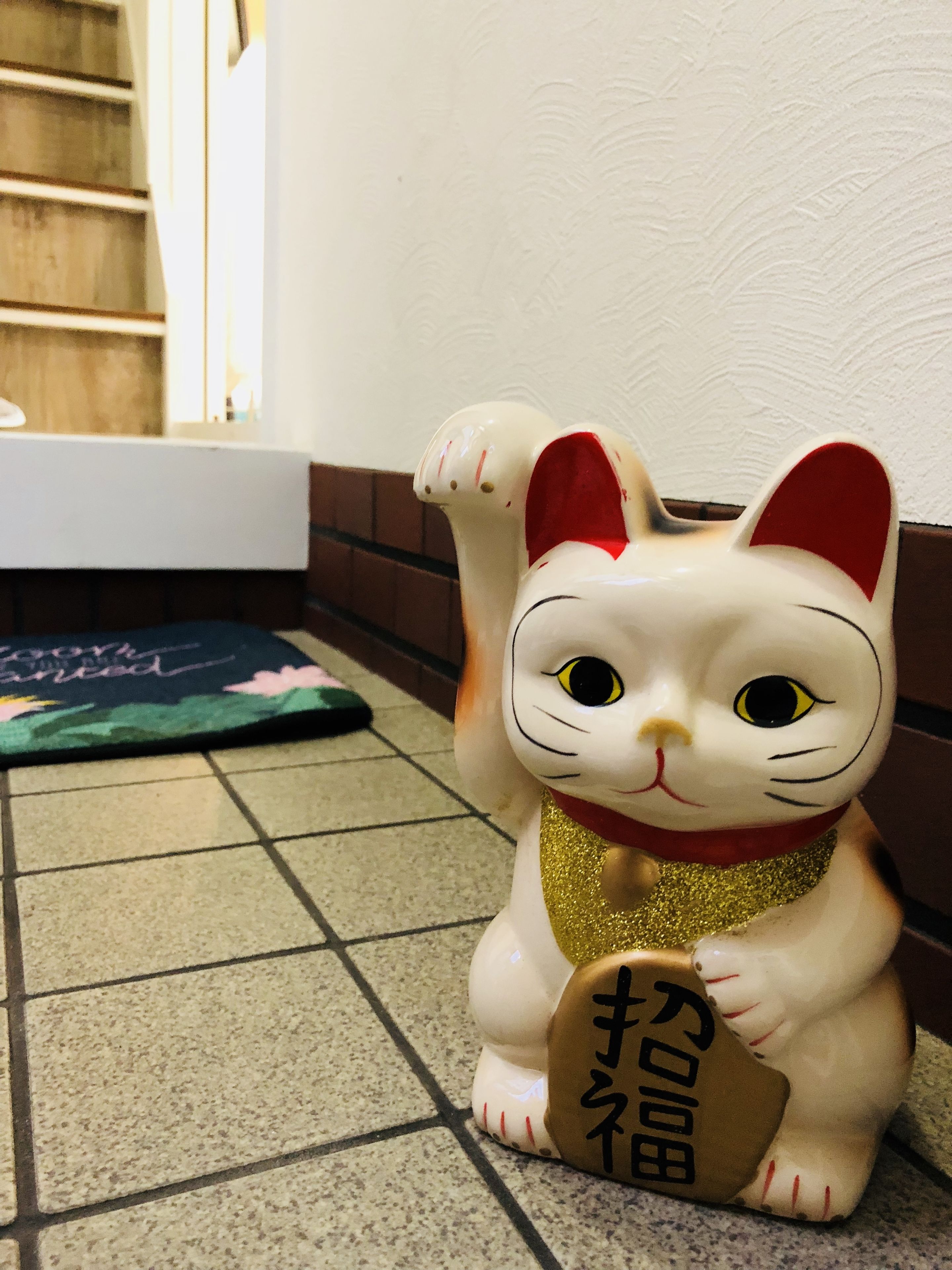 A ceramic cat figurine with a red collar and a gold bell, sitting on a tiled floor.