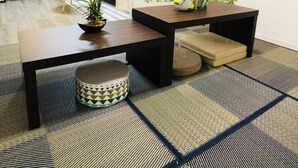 Een moderne, goed verlichte kamer met een houten tafel, een bank en een kamerkleed met een patroon. Op de tafel staan planten in potten en een muursticker met een boom en het woord 'SPRING'.