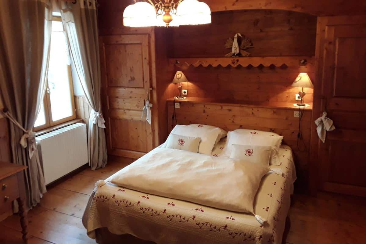 Une pièce en bois avec un lit, deux lampes de chevet, un radiateur et une fenêtre avec des rideaux.