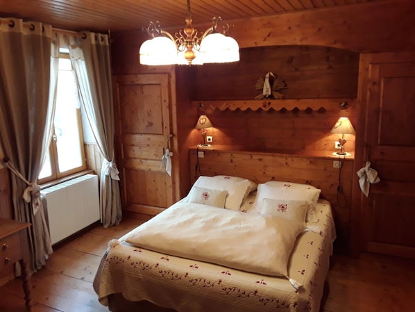 A wooden room with a bed, two bedside lamps, a radiator, and a window with curtains. - Chambre d'hote le Bellevue (Giron)