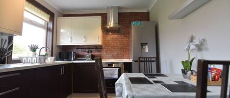 A modern kitchen with dark wooden cabinets, a brick wall, a stainless steel refrigerator, and a dining area with a white tablecloth.