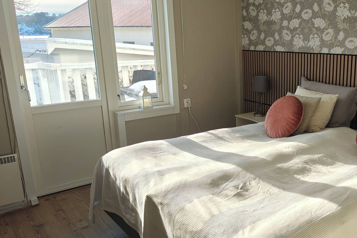 Une chambre à coucher avec un lit, une table de chevet, une lampe et une fenêtre donnant sur des immeubles.