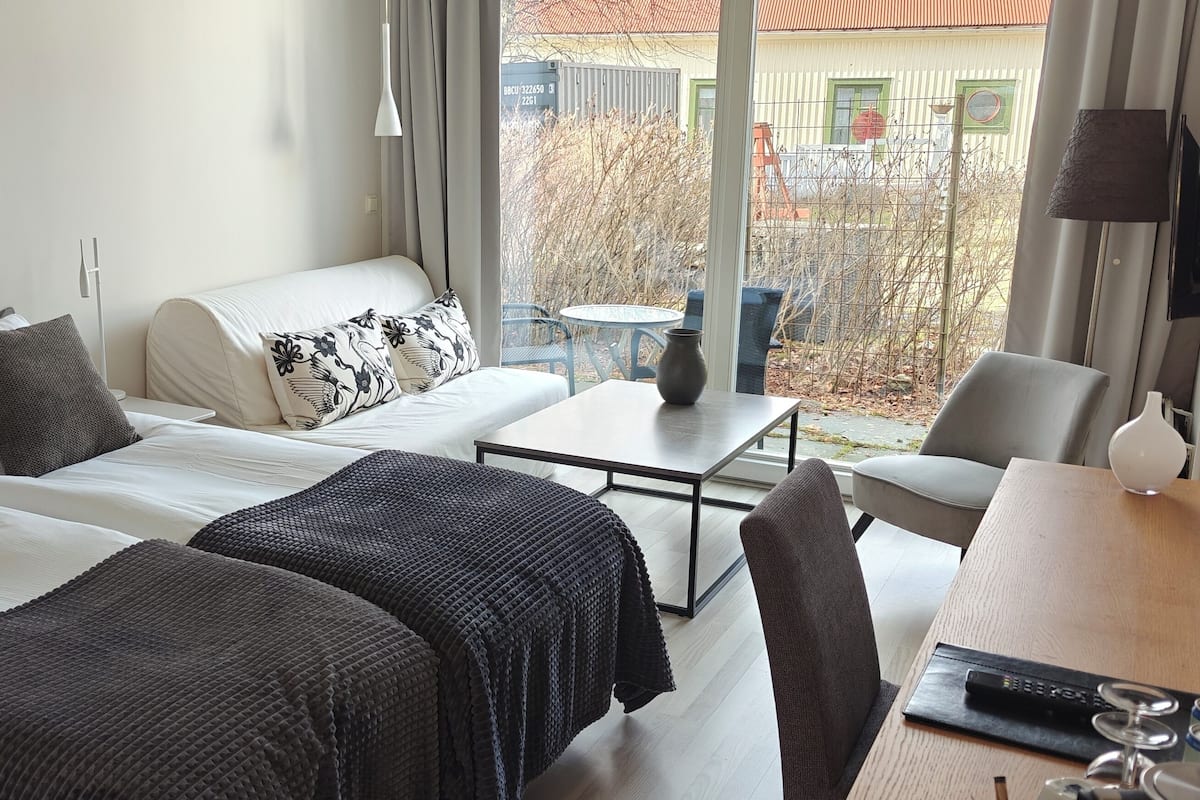 Une chambre d’hôtel avec deux lits, un canapé, une petite table et une chaise. De grandes fenêtres offrent une vue sur l’extérieur.