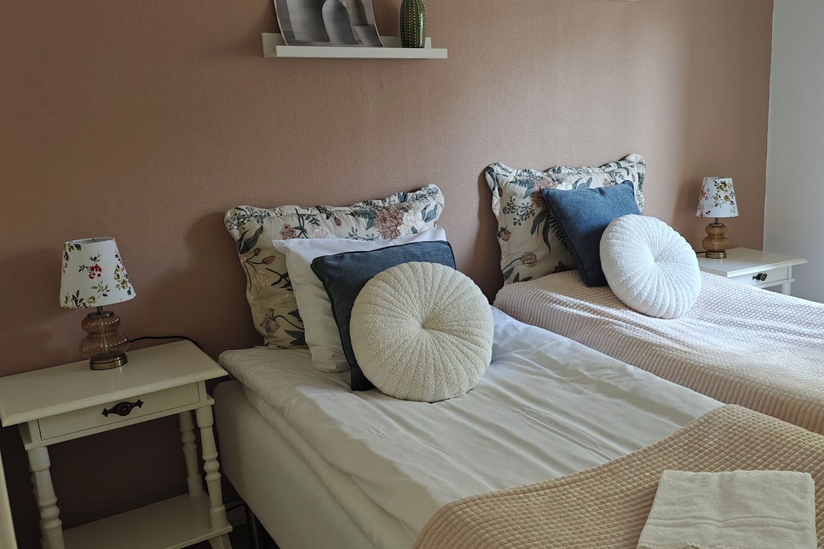 Une chambre avec deux lits, une table de chevet avec une lampe, une petite table et une fenêtre avec des rideaux.