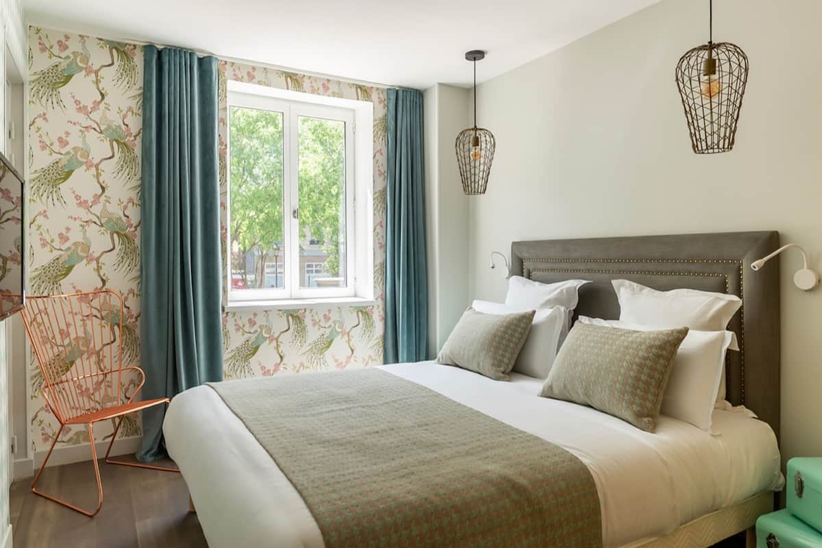 A bedroom with a large bed, a chair, a window with curtains, and two hanging light fixtures.