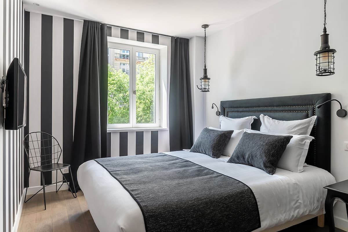 A modern bedroom with a large bed, black and white striped curtains, a view of greenery outside, and a black metal chair.