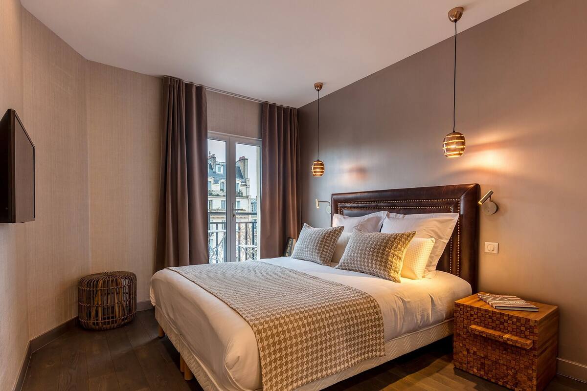 A modern bedroom with a large bed, a bedside table, a small round ottoman, and a view of buildings through a window.