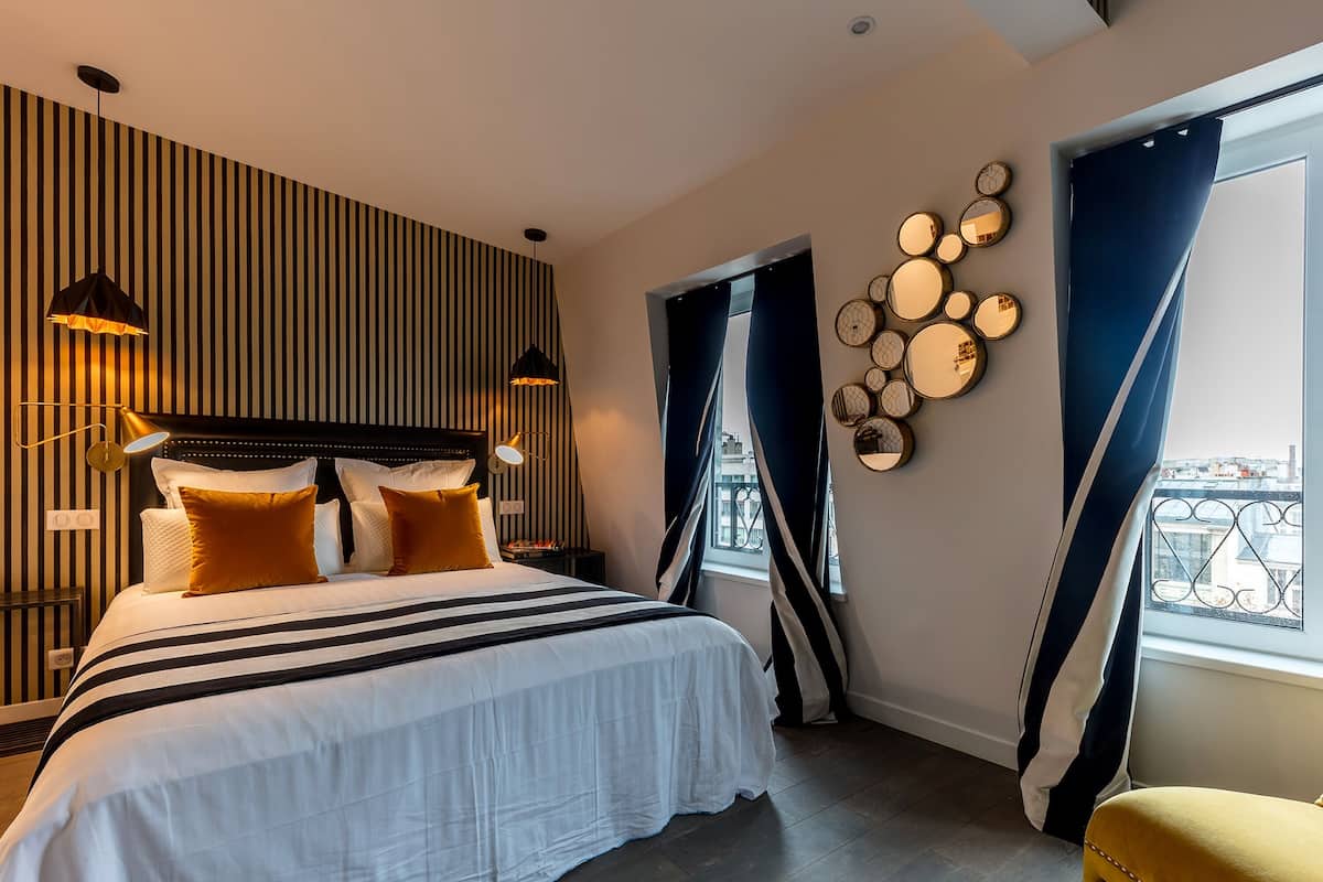 A modern hotel room with a large bed, striped wall paneling, a decorative wall art piece, and a view of the city through a window with curtains.