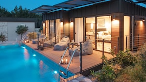 Area piscina esterna moderna con terrazza in legno, posti a sedere e camera da letto con vetrate e bagno visibile.