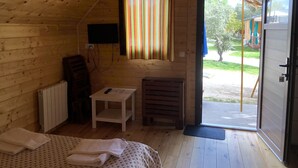 Een gezellig houten huisje met een bed, een klein tafeltje, een stoel, een radiator, een aan de muur bevestigde televisie en een raam met een gestreept gordijn.