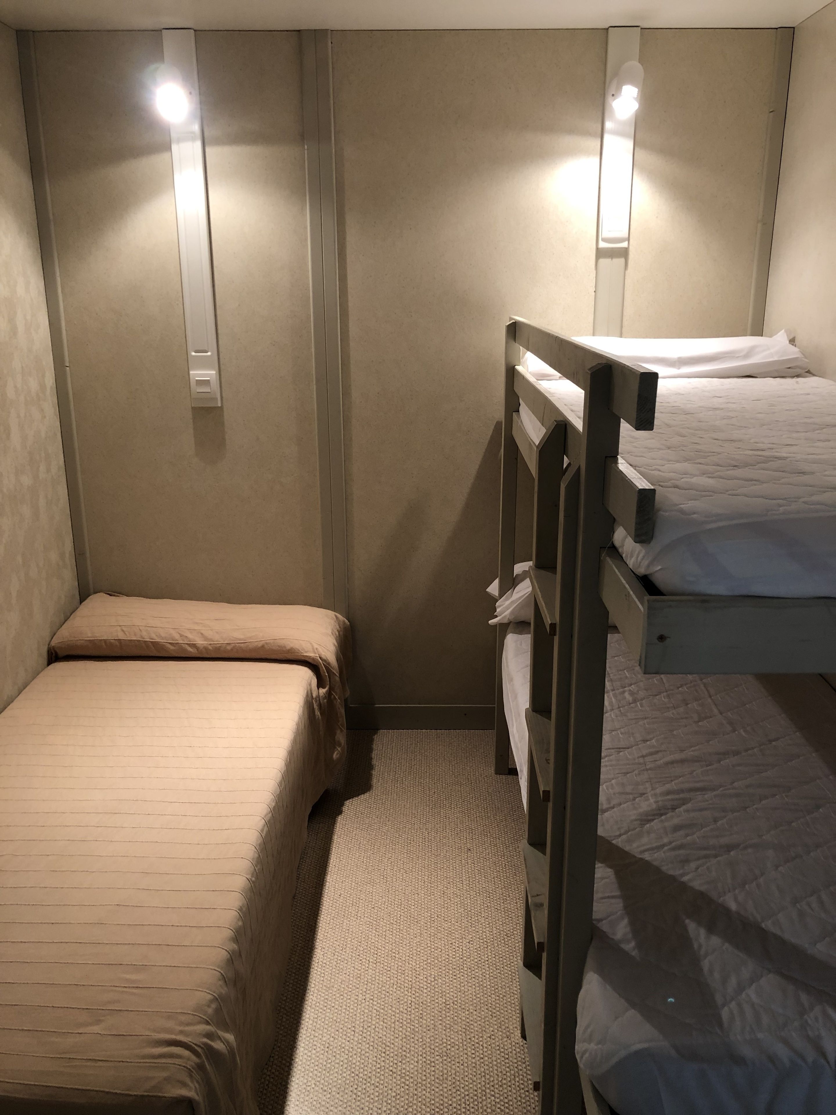 Una habitación pequeña, con poca luz, que tiene dos literas, una cama individual y una lámpara fijada a la pared.