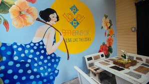 Une fresque représentant une femme portant une robe à pois, un cercle jaune avec du texte, et une table sur laquelle se trouve une plante.