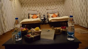A small room with two beds, a table with water bottles, a bowl of fruit, and a sign. - TSI Kumbh Mela Camp (Prayagraj)