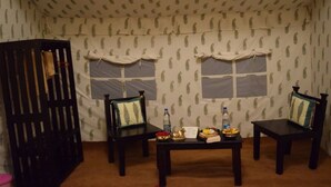 A room with two wooden chairs, a table with a bottle and a bowl, and a patterned wall. - TSI Kumbh Mela Camp (Prayagraj)