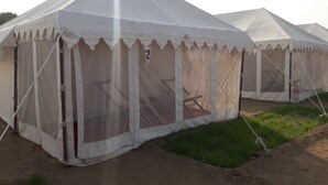 A row of white tents with a peaked roof and a mesh door, set on a dirt ground with grass in the foreground. - TSI Kumbh Mela Camp (Prayagraj)