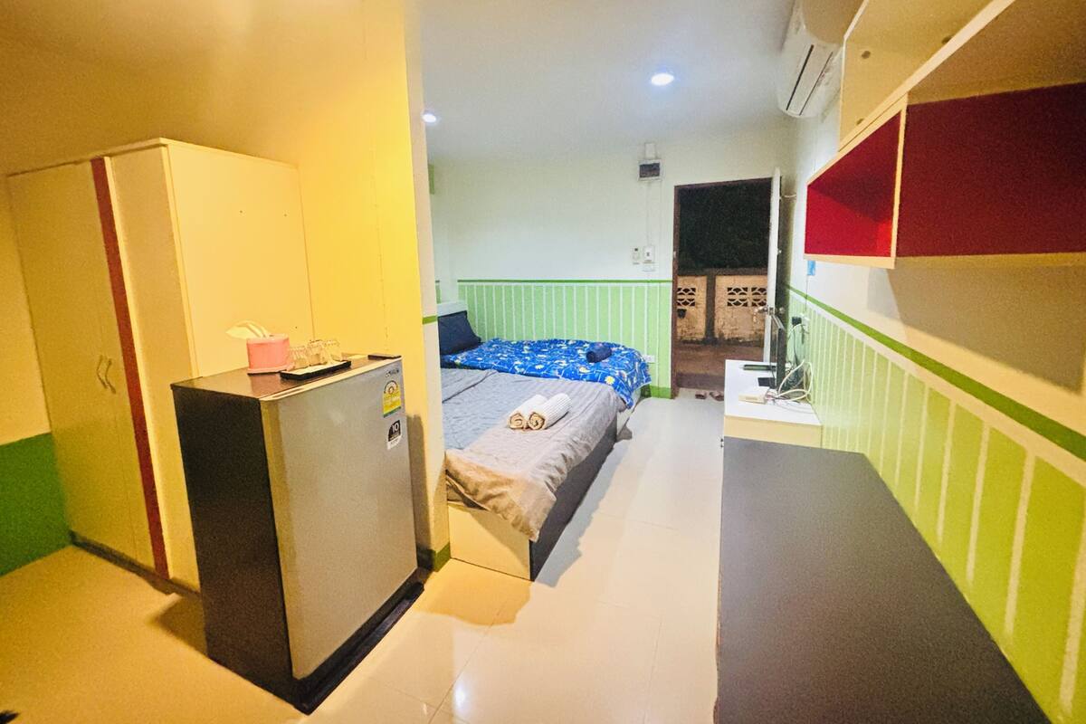 A compact hotel room with a bed, a small desk, a wardrobe, and a kitchenette.